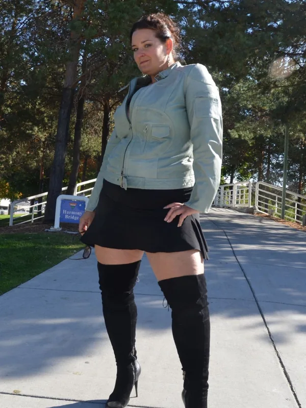 Femme debout sur un trottoir en plein air, portant une veste claire, une jupe noire courte et des bottes hautes, regardant l'objectif.