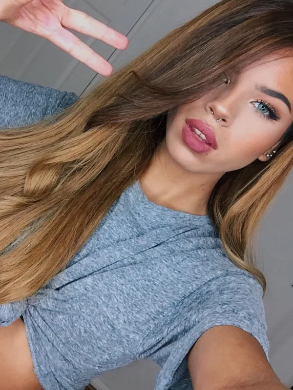 Portrait en selfie d'une jeune femme aux cheveux longs bruns et blonds, portant un haut gris noué, faisant un signe de victoire.