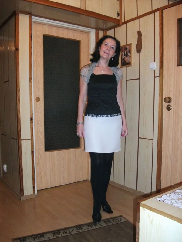 Femme debout en pied dans un intérieur en bois, portant une jupe blanche, un haut noir et des collants noirs.