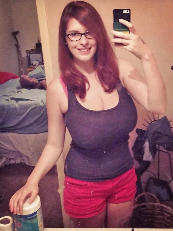 Femme aux cheveux roux et lunettes prenant un selfie dans un miroir, vêtue d'un débardeur gris et de shorts roses, dans une chambre.