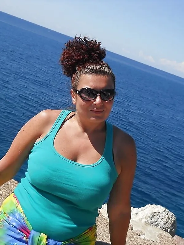Femme portant un débardeur turquoise et une jupe colorée, posant sur un rocher devant la mer sous un ciel bleu.