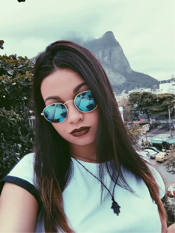 Portrait d'une femme aux cheveux longs portant des lunettes de soleil rondes, devant une vue urbaine avec une colline rocheuse.