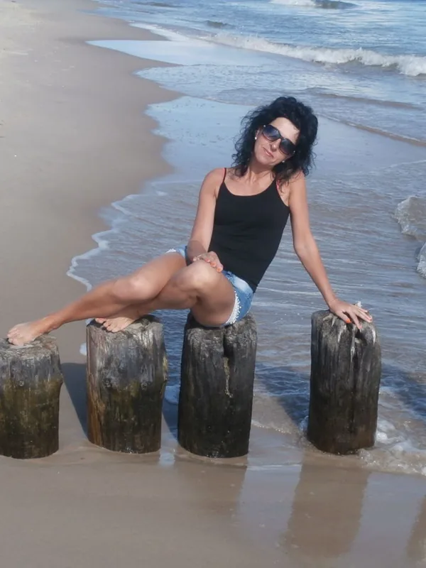 Femme assise sur des poteaux en bois au bord de la mer, portant un haut noir et un short en jean, sous un soleil vif.