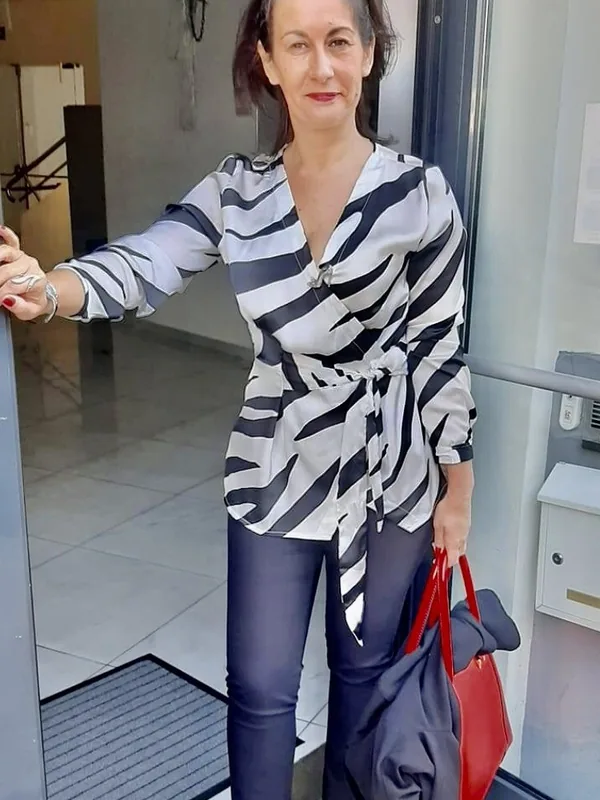 Femme debout dans un hall, portant une blouse zébrée, des pantalons sombres et des chaussures rouges, tenant un sac rouge.