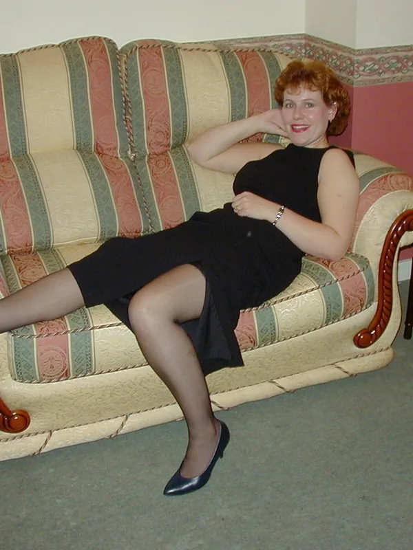 Femme aux cheveux roux assise sur un canapé à rayures, portant une robe noire et des chaussures à talons.