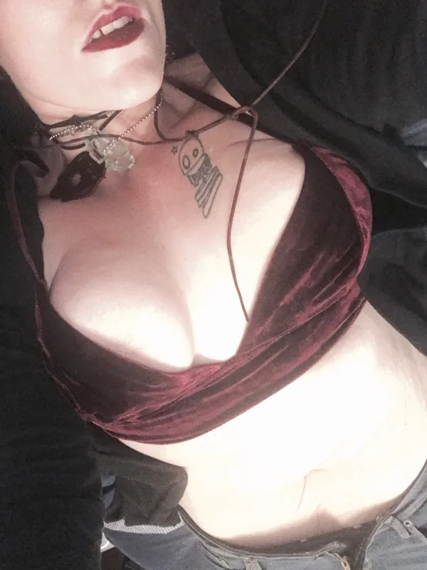 Portrait rapproché d'une personne portant un haut en velours bordeaux, une veste noire et un collier, avec un tatouage visible sur le torse.