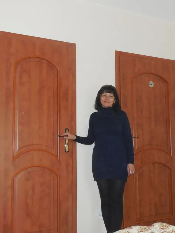 Femme debout entre deux portes en bois dans une pièce blanche, vêtue d'un pull bleu et de leggings sombres.
