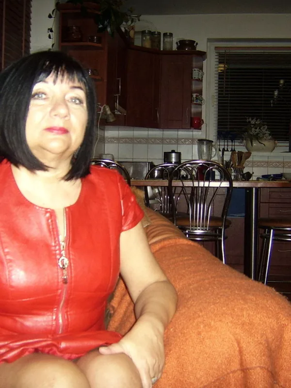Femme aux cheveux noirs portant une robe rouge assise sur un canapé dans un salon avec cuisine en arrière-plan.
