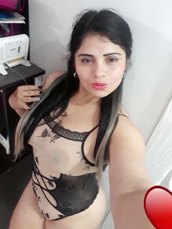 Portrait en selfie d'une femme aux cheveux longs portant un body transparent à motifs floraux, regardant l'objectif dans un cadre intérieur.