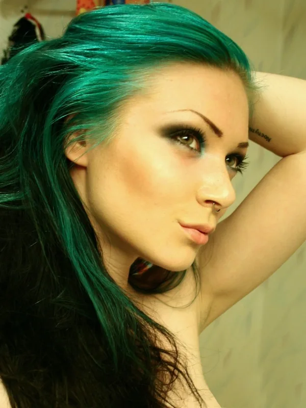 Portrait d'une jeune femme aux cheveux verts, maquillée, regardant vers l'objectif, main derrière la tête, peau nue au niveau des épaules.