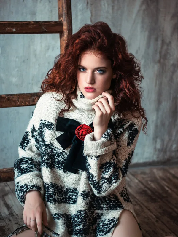 Femme aux cheveux roux portant un pull tricoté blanc et noir avec une rose rouge, assise sur une chaise en bois.